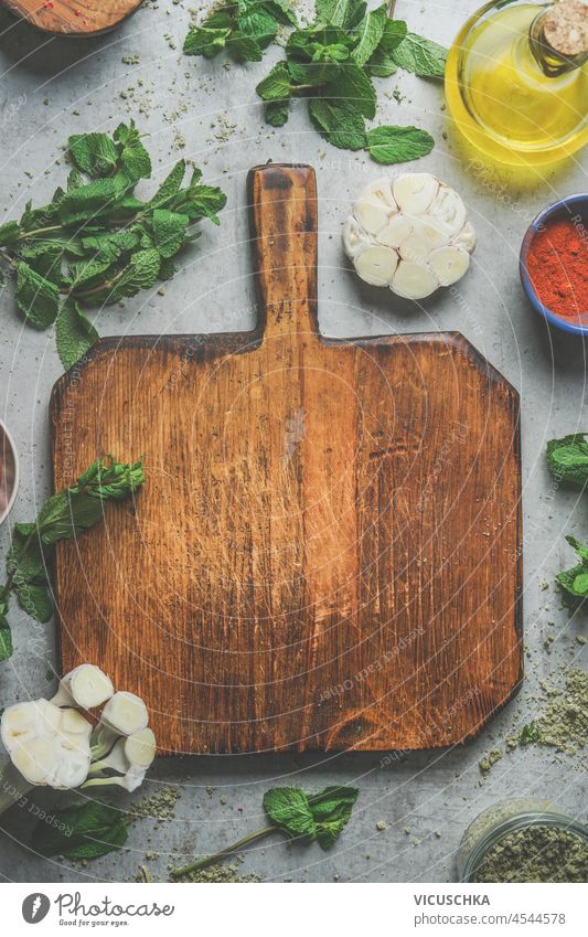 Food background with empty wooden cutting board, herbs, garlic, olive oil, spices on grey concrete kitchen table. Cooking with fresh, flavorful ingredients. Top view with copy space.