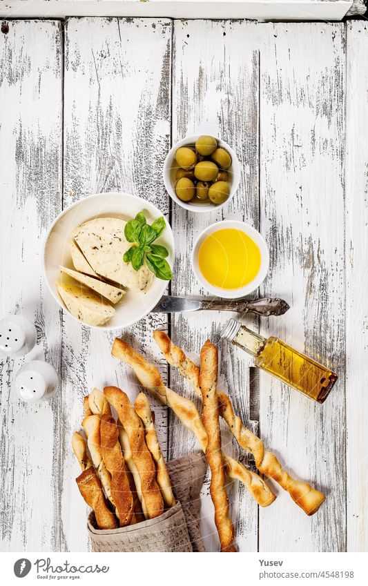 Crispy grissini breadsticks. Traditional Italian wheat bread with garlic, cheese and sesame seeds. Food still life on a white rustic background. Olive oil, goat cheese and green olives. Top view