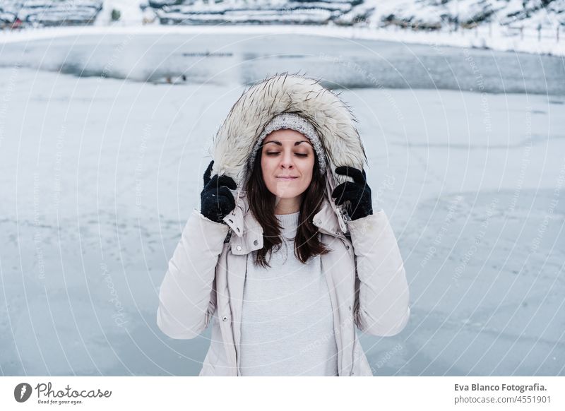 portrait of relaxed beautiful caucasian woman with eyes closed holding hood coat and standing in front of frozen lake in city. Lifestyle during winter in city