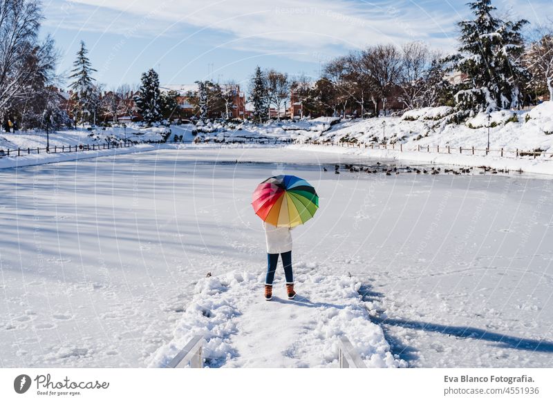 back view of woman holding colorful umbrella standing on snowy pier by frozen lake during winter on sunny day. winter lifestyle in park frost dock city backpack