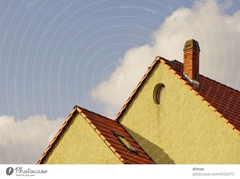 Two dull yellow house gables with a chimney, a skylight, an oval gable window and red roof tiles in front of a blue sky and decorative clouds / live pediment