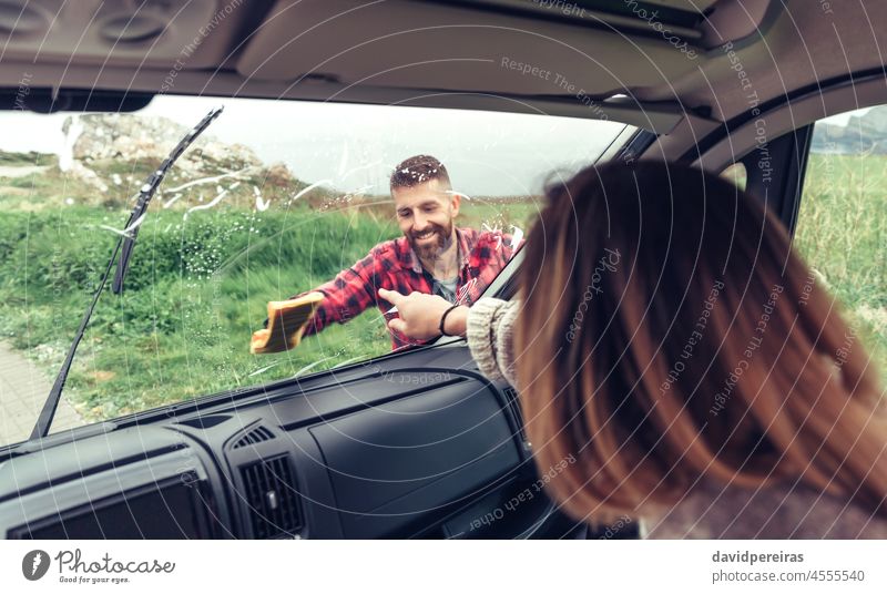 Man cleaning van windshield while woman pointing stain smiling rinsing glass cloth view from inside through the glass outdoors careful owner camper
