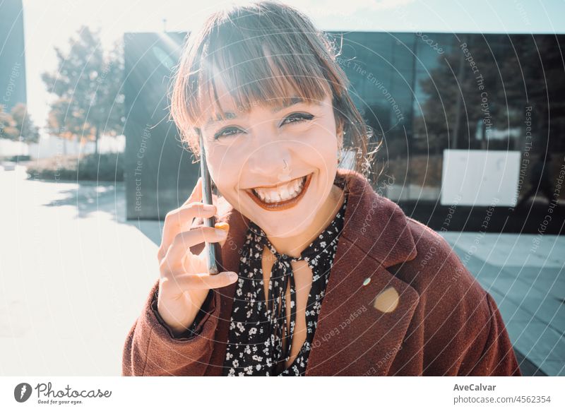 Young beautiful business woman in red coat using mobile phone and looking away standing on the financial street. Finances concept, young business. Smiling to camera during a sunny day