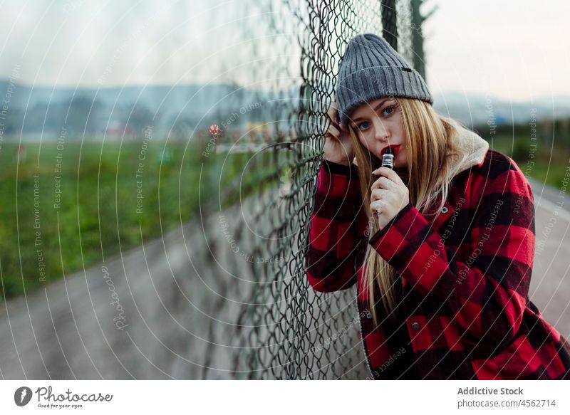Young woman with wool hat leaning on the fence and smoking portrait young vaping alternative looking at camera attractive vaporizer beautiful blonde blow
