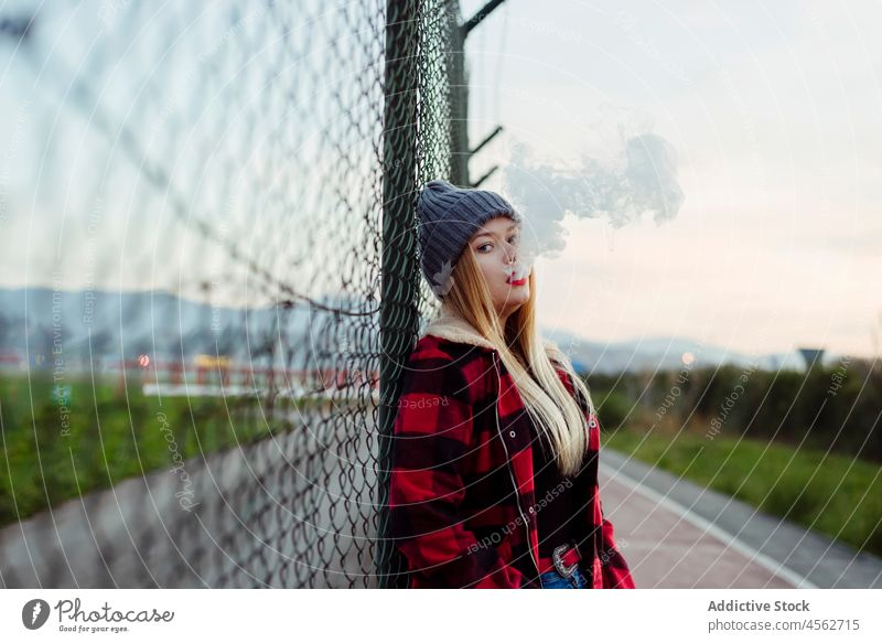 Young woman with wool hat leaning on the fence and smoking young vaping alternative looking at camera attractive beautiful blonde blow caucasian cigarette city