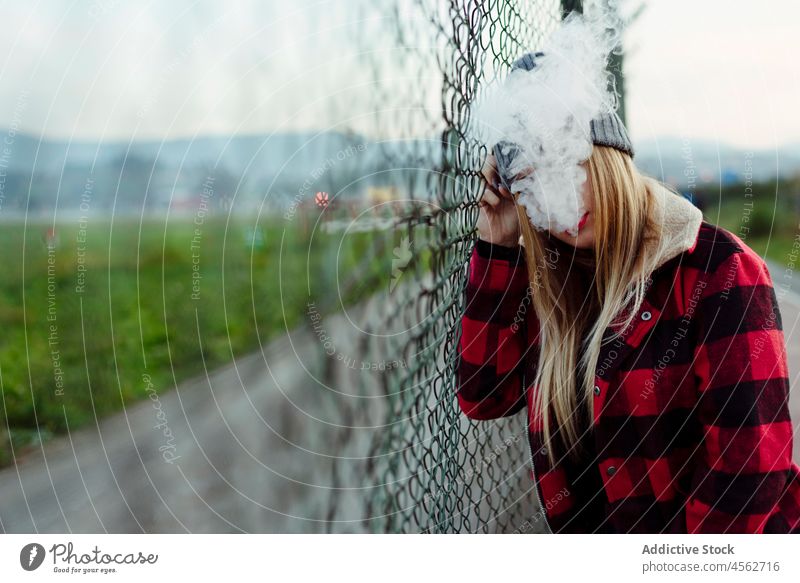 Young woman with wool hat leaning on the fence and smoking young adult vaping alternative faceless attractive beautiful anonymous blonde blow caucasian