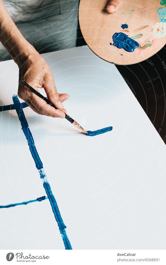 Close-up of Female senior old Artist Hand, Holding Paint Brush and Drawing Painting with blue Paint. Colorful, Emotional Oil Painting. Contemporary Painter Creating Modern Abstract Piece of Fine Art