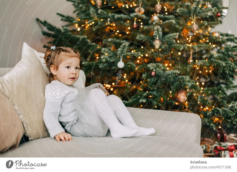A pretty girl in a hat is sitting on the sofa near a Christmas tree, dressed up for the holiday, the child is smiling and rejoicing at the gift little girl