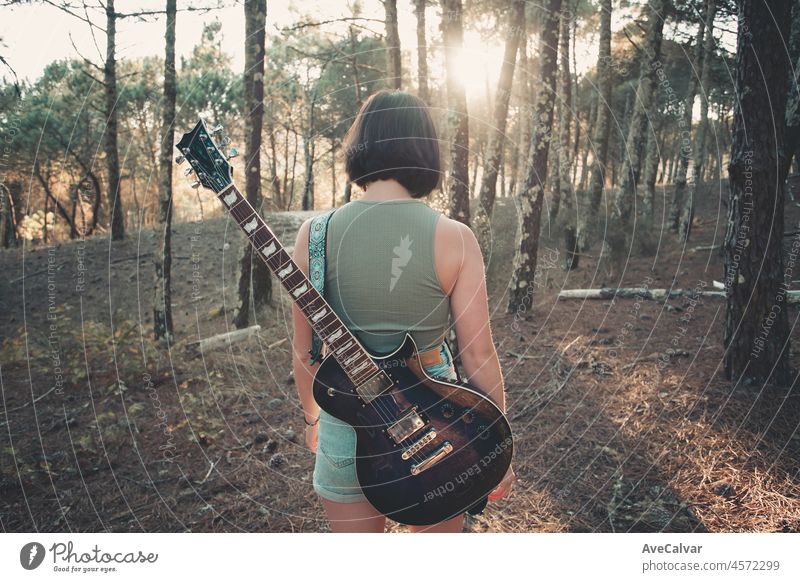 Back view of a young hipster woman playing the guitar smiling outside the forest park of the city. Having fun learning a new skill, music play seasonal style. Young short hair girl. Copy space