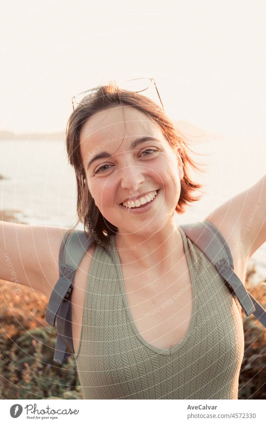 Smiling portrait young woman walks on the path of rocks on the shore and looks at the evening sea.Young hipster girl doing some hiking on the shore, traveling and low budget travel concepts,copy space