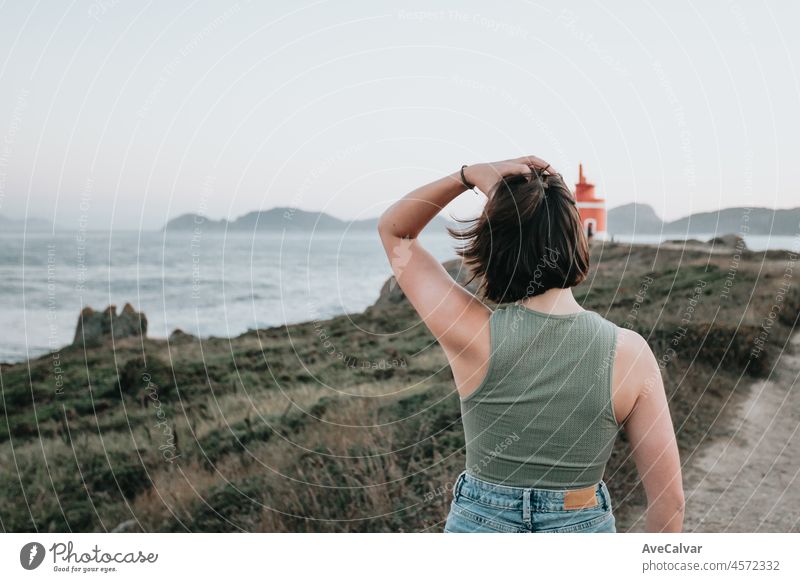 Back view sunset young woman walks on the path of rocks on the shore and looks at the evening sea.Young hipster girl doing some hiking on the shore, traveling and low budget travel concepts,copy space