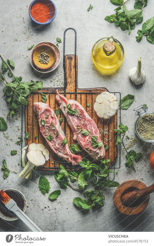 Raw meat with ingredients on wooden cutting board at  kitchen table background with fresh cooking ingredients: olive oil, garlic, herbs and spices. Top view.