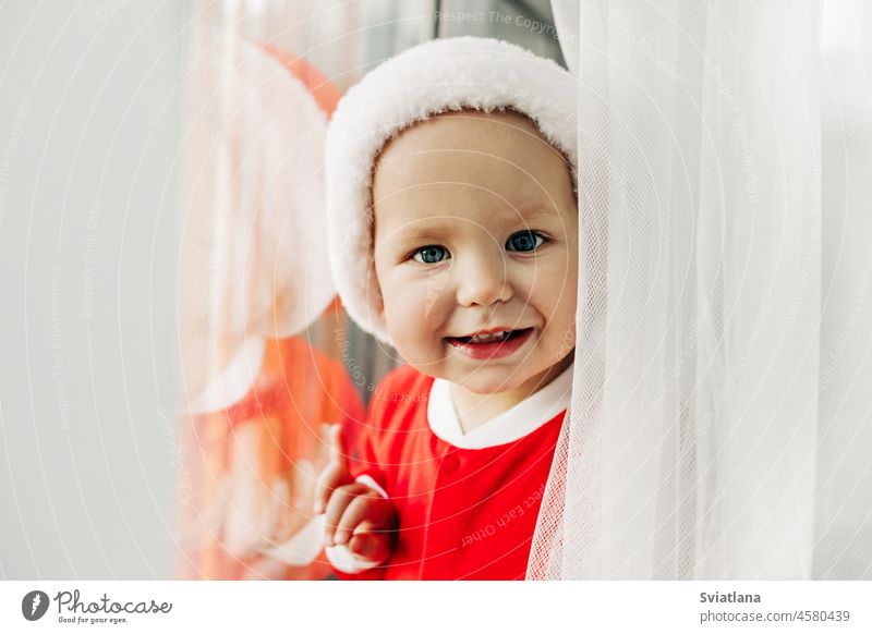 Portrait of a charming baby in a Santa Claus costume at the window. Close-up. The concept of Christmas and New Year christmas santa waiting hat happy happiness