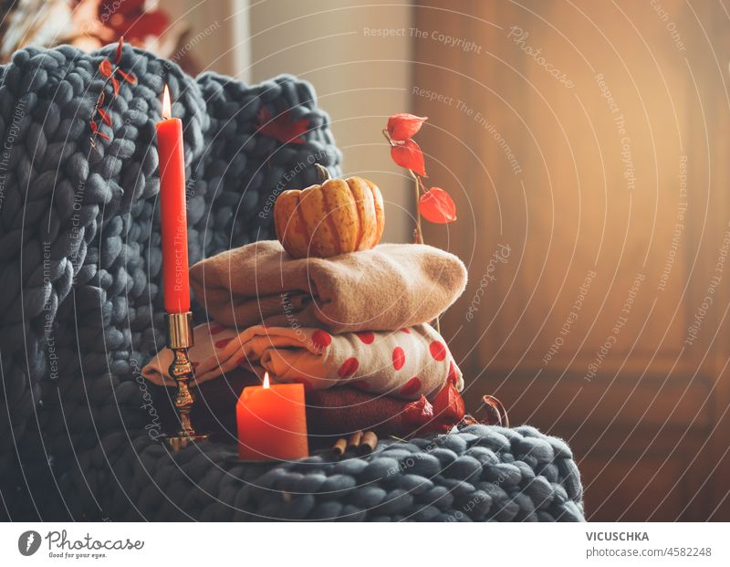 Cozy autumn still life with knitted sweaters, candles, grey wool blanket, candles and autumn leaves at interior background. Warm clothes for autumn season. Front view.