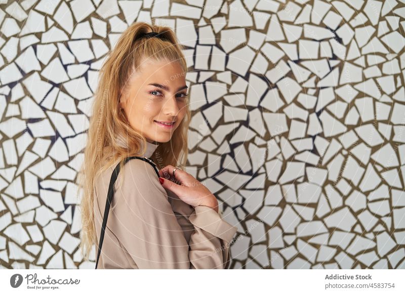 Young woman with backpack standing at mosaic wall trendy urban street walk outfit city blond leather casual calm daytime sunlight way building walkway stroll