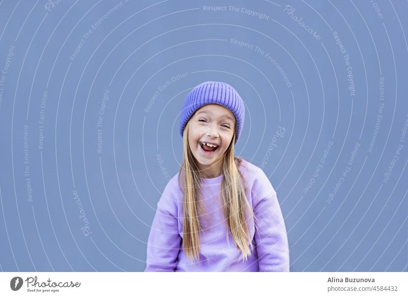 Cute little caucasian girl eight years old with blonde hair smiling outdoor. Kid wearing stylish shirt and knitted hat violet color. Trendy color of the 2022 year very peri