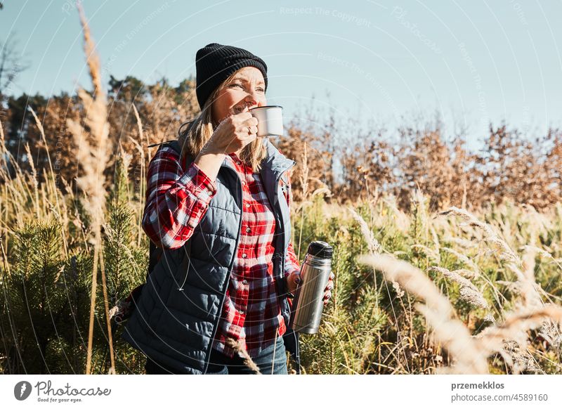 Woman taking break and relaxing with cup of coffee during summer trip. Woman standing on trail and looking away. Woman with backpack hiking through tall grass along path in mountains