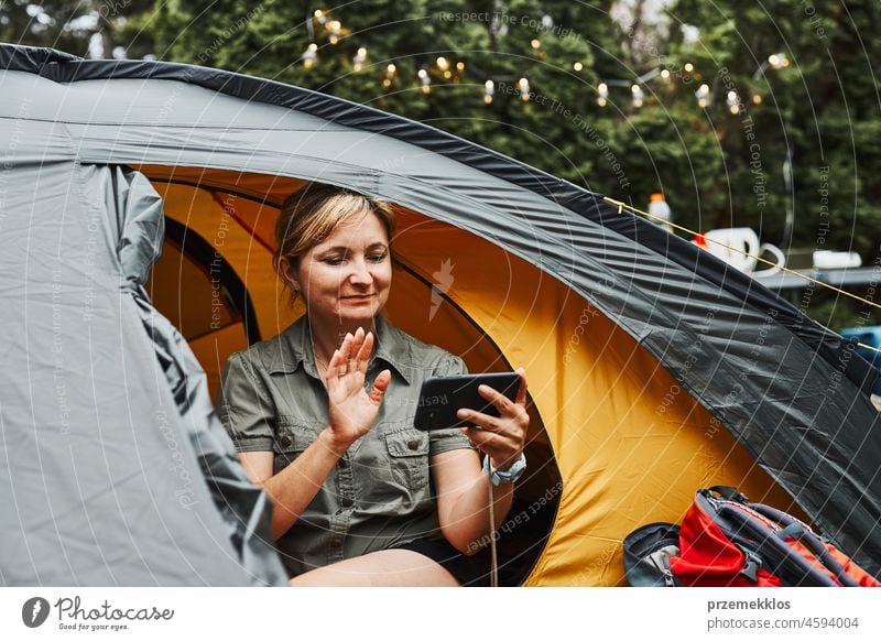 Female having video call with friends using smartphone while sitting in tent at camping. Woman relaxing in tent during summer vacation trip adventure campsite