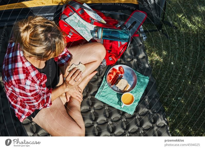 Woman having breakfast sitting by tent at camping. Female actively spending summer vacations close to nature outdoors trip adventure campsite traveling food