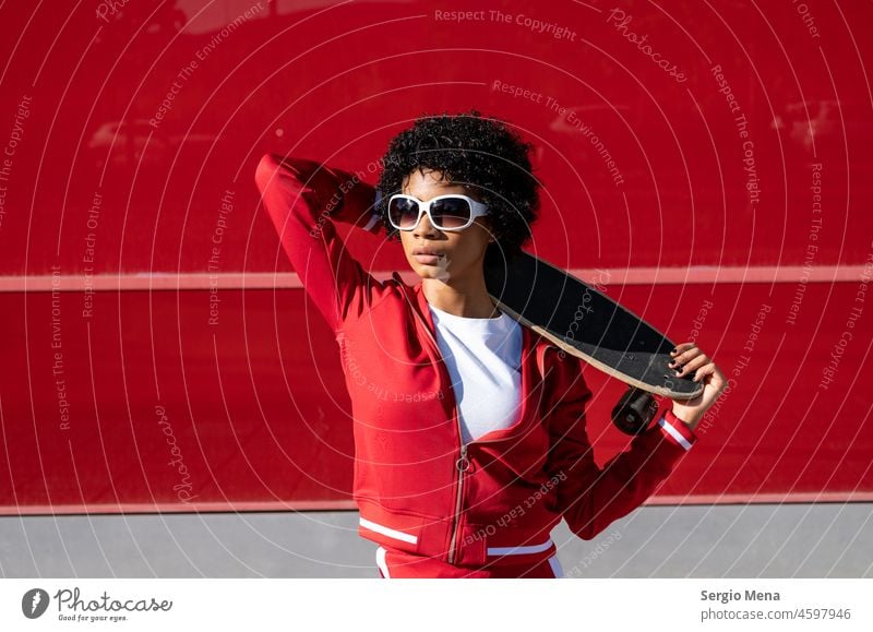 African American woman with short hair, sunglasses and red sportswear with her skateboard female Spain clothes wall background standing holding curly hair