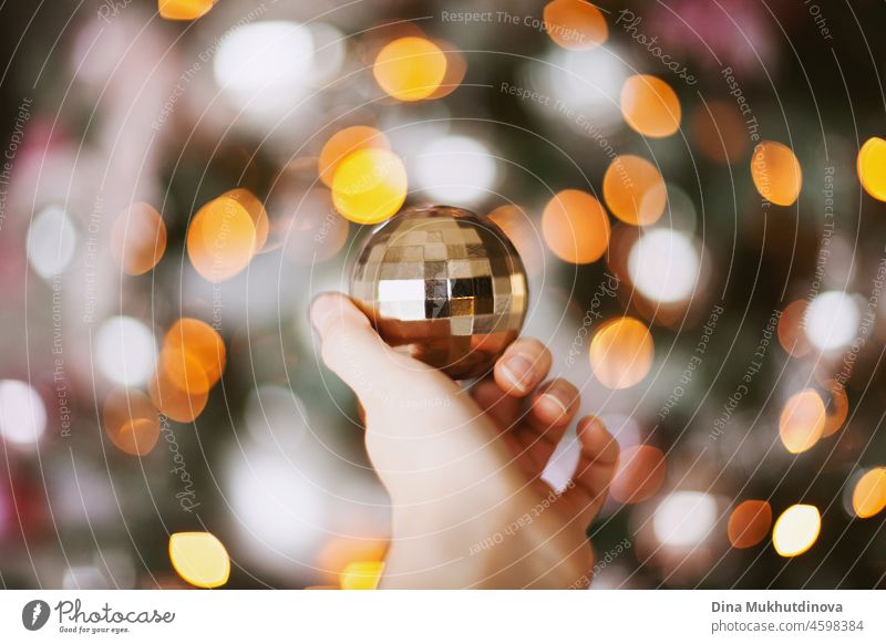 Hand holding a shiny gold christmas ornament with christmas tree lights bokeh as background shine sphere bauble festive merry year celebration seasonal ball