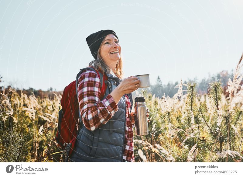 Smiling woman relaxing and enjoying the coffee during vacation trip. Woman standing on trail and looking away holding cup of coffee and thermos flask. Woman with backpack hiking through tall grass along path in mountains