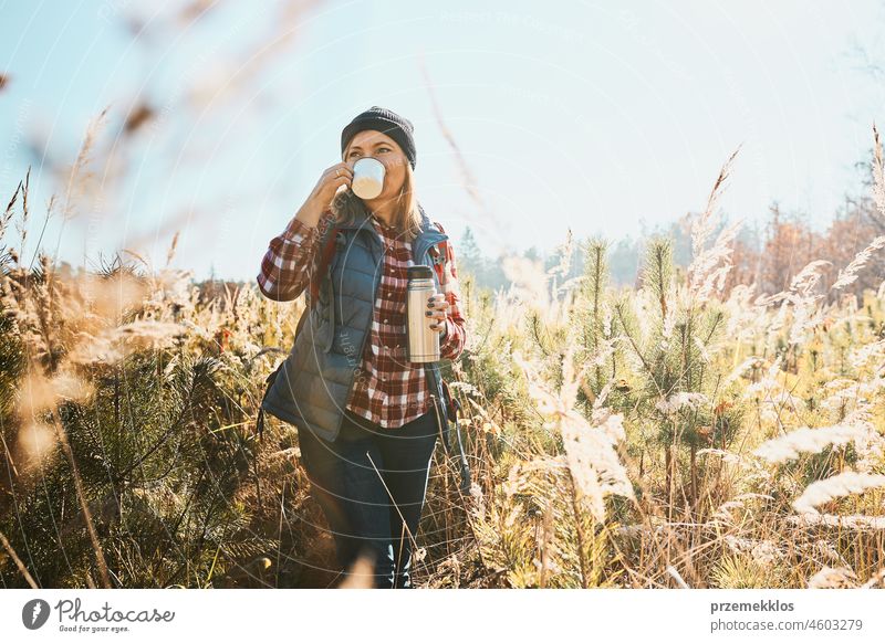 Woman enjoying the coffee in bright warm sunlight during vacation trip. Woman standing in tall grass and looking away holding cup of coffee and thermos flask. Woman with backpack hiking along path in mountains