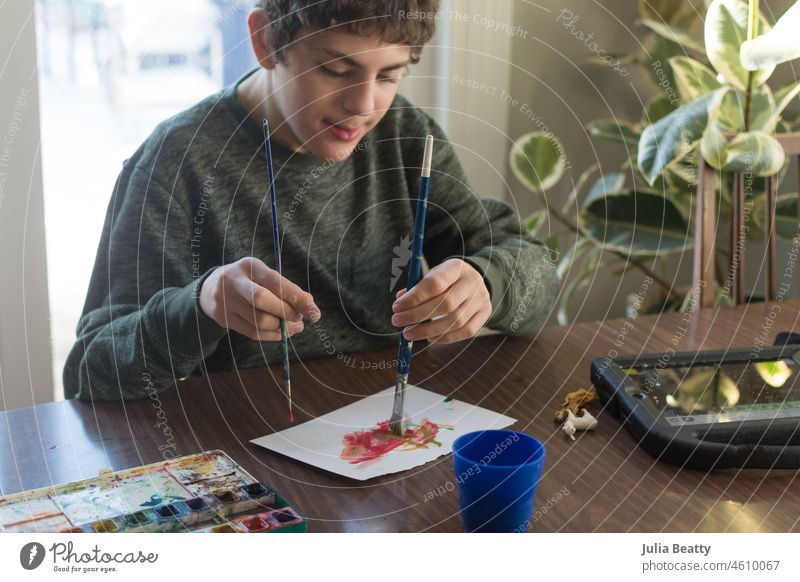 Boy with special needs water color painting with both hands at the same time; AAC communication device sits nearby low muscle tone autism grasp hold paint brush