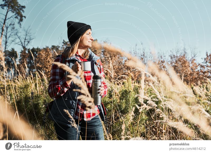 Woman taking break and relaxing with cup of coffee during summer trip. Woman standing on trail and looking away. Female with backpack hiking through tall grass along path in mountains