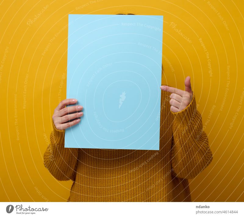 woman in an orange sweater holds a blank sheet of paper on a yellow background. Place for an inscription, advertisement, information person poster holding empty