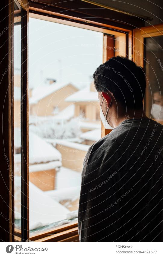 Young man with mask in quarantine looking at the snowfall through the window of his house men boy young confined confinement medical mask person virus disease