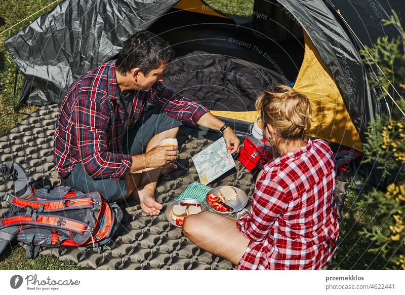 Couple having breakfast and planning their day sitting by tent at camping. People actively spending summer vacations close to nature outdoors trip adventure
