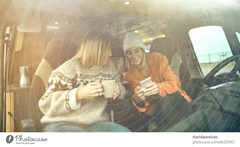Women laughing looking mobile sitting in the front seat of a camper van happy women fun drinking coffee copy space through the windshield winter woman
