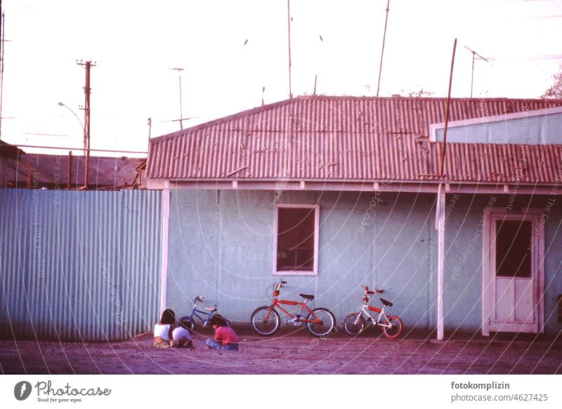 Three children with three wheels sitting on the ground in front of a house in South America Infancy Friends free time Playing Children's game Playground Bicycle