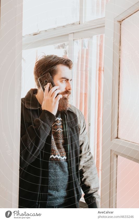 Handsome young bearded man making calls and checking his phone on the window of his house. Looking outside the window while chatting on his phone. Long beard and hipster style