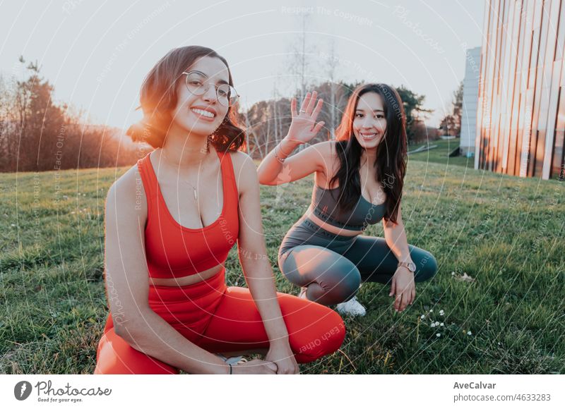 Two young african females resting while squatting at the grass during a colorful sunset, portrait one in focus. Training at the park after work, workout at the city, having fun together doing exercise