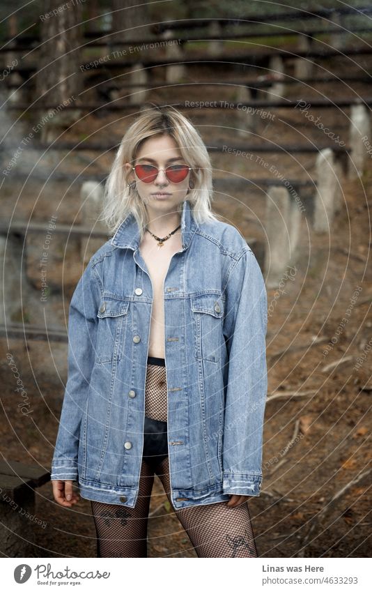 A gorgeous blonde girl is standing in front of a camera. With her blue jeans jacket, red sunglasses, inked and pierced body. Some smoke is surrounding her beautifully.