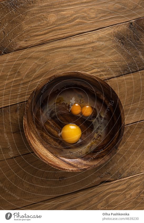 Wooden round bowl with broken raw fresh chicken eggs. Double yolk egg and usual egg look like funny face with eyes and nose. Natural brown wooden table background. Directly above view. Copy space.