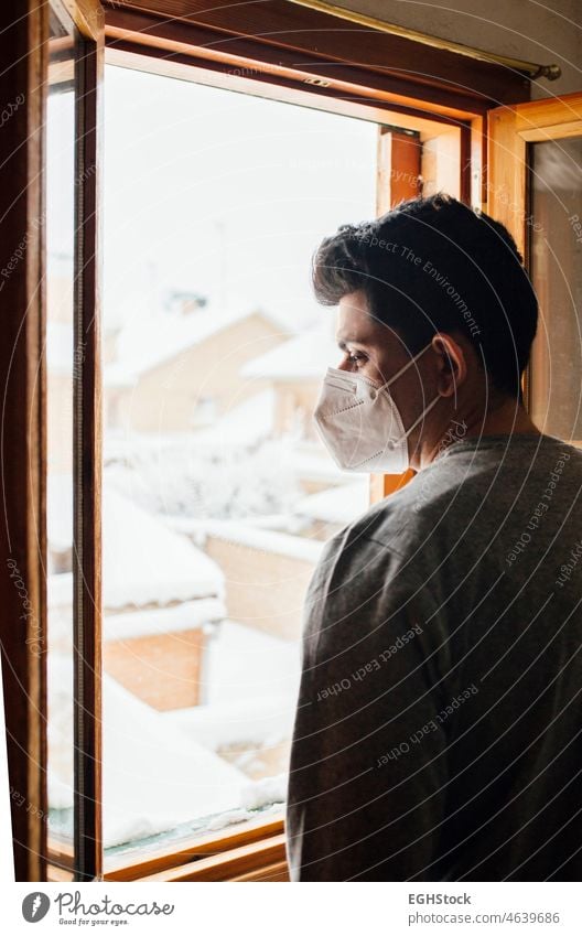 Young man with mask in quarantine looking at the snowfall through the window of his house alone anxiety boy confined confinement contemplation corona