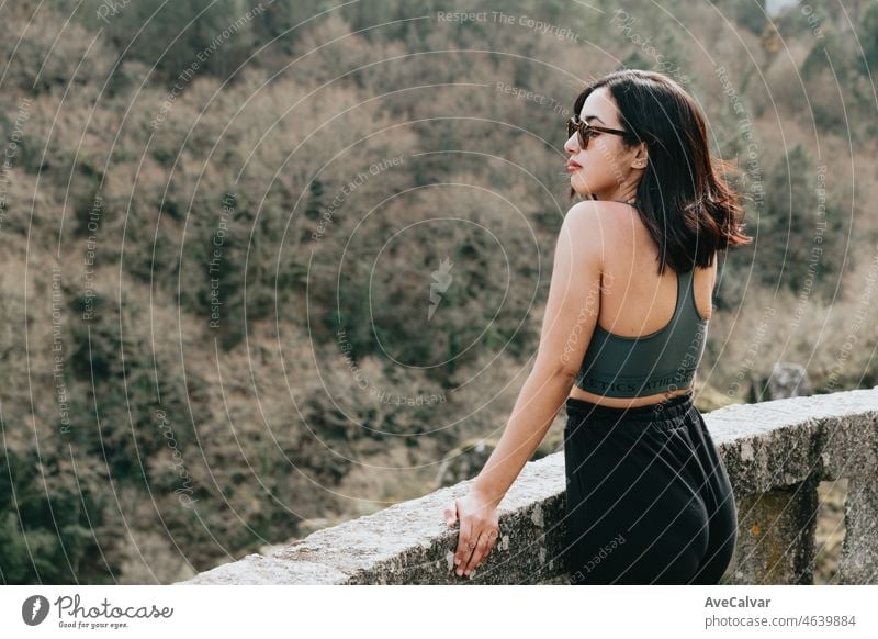 A young woman traveler in sporty clothes and sunglasses with looking from a cliff in the forest, admiring the views of the wildlife. Healthy life at the nature young people concept.