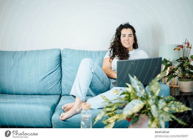 Young freelancer latina woman working on computer while sitting on the comfortable sofa at home on modern trendy clothes. Concept of remote work from home, young people online works