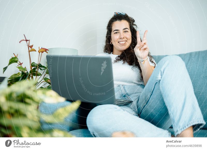 Young freelancer latina woman working on computer while sitting on the comfortable sofa at home smiling to camera doing victory sign on modern trendy clothes. Concept of remote work from home