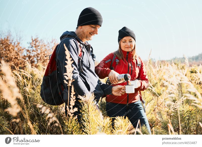 Couple relaxing and enjoying the coffee during vacation trip. People standing on trail pouring coffee from thermos flask. Couple with backpacks hiking through tall grass along path in mountains. Active leisure time close to nature