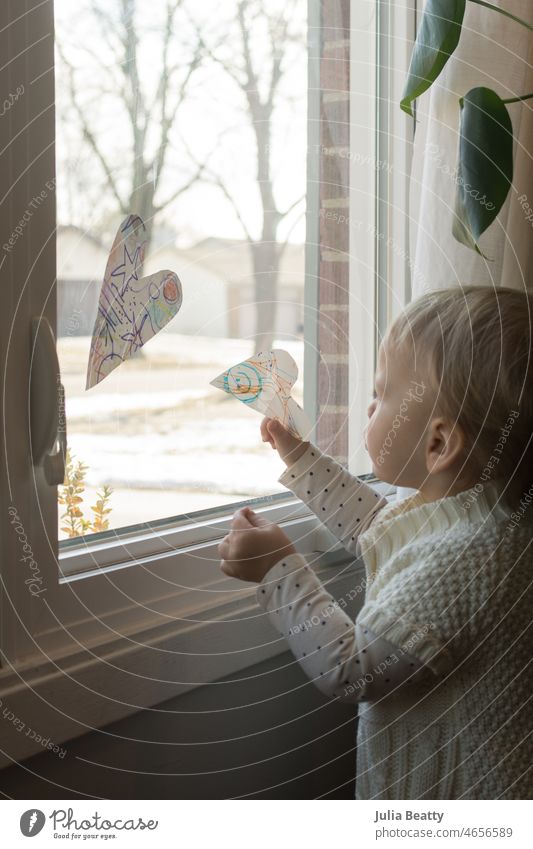 Toddler hangs handmade paper hearts in the window of her home; DIY Valentine's Day decorations valentine's day diy homemade craft arts and crafts toddler
