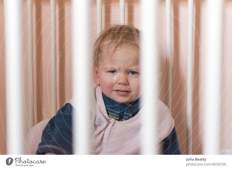 Toddler wakes from a nap with a questioning expression; baby in a wearable blanket in a peach and white nursery child toddler wake up sleep bedtime routine