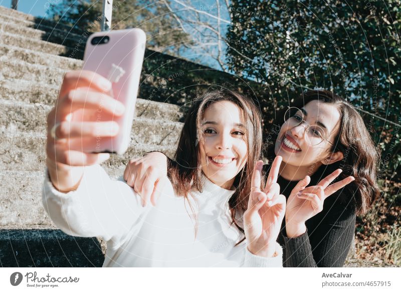Two young students sitting outdoors make selfie by phone, university building on background and looking at smart phone. Young woman celebrating study on trendy clothes super smile, multi racial