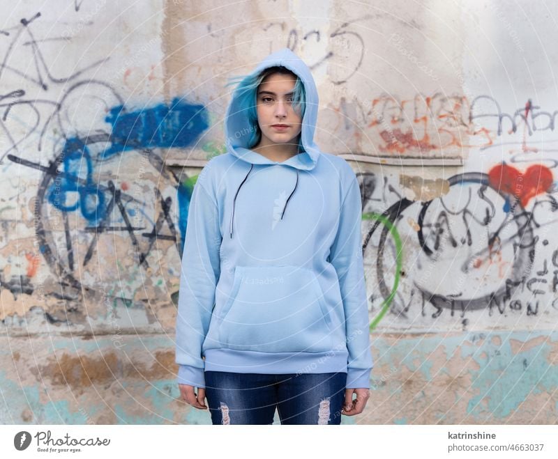 Blue haired Teenage girl in light blue oversize hoodie pointing against graffiti wall Teenager mockup jeans blue haired teen girl outdoors urban modern hipster