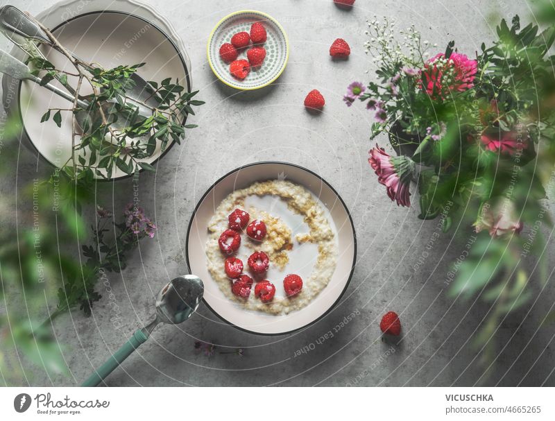 Homemade porridge bowl with milk raspberries at grey concrete kitchen table homemade bowls cutlery beautiful summer flowers eating healthy breakfast at home