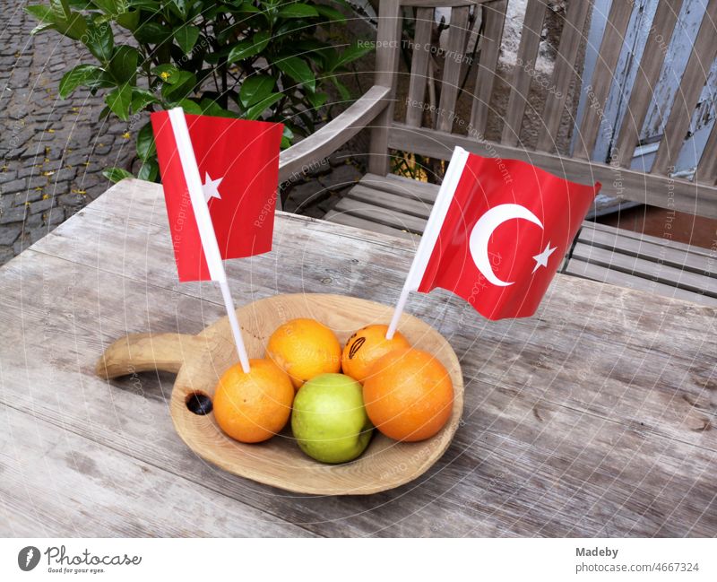 Fresh oranges and green apple with red Turkish flags in a decorative wooden bowl on a rustic wooden table with wooden bench in the alleys of the old town of Alacati near Izmir in Turkey