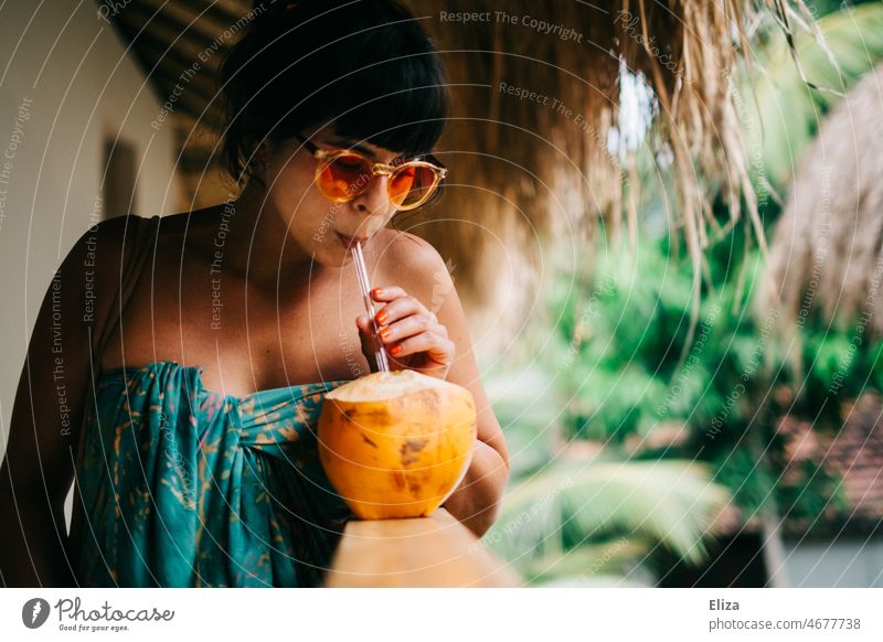 Smiling woman with sunglasses wrapped in colorful bath towel drinking from a King coconut with straw in hand. Vacation in the tropics. Coconut Drinking coconut